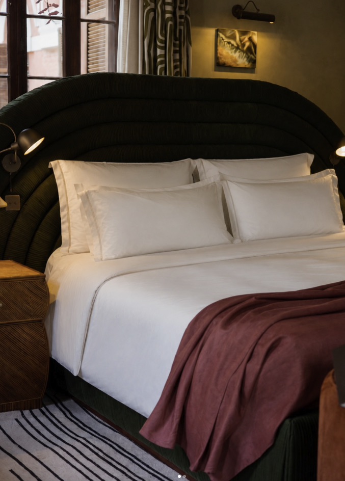 Cama arrumada com lençóis brancos e manta em tom vinho, cabeceira curva escura